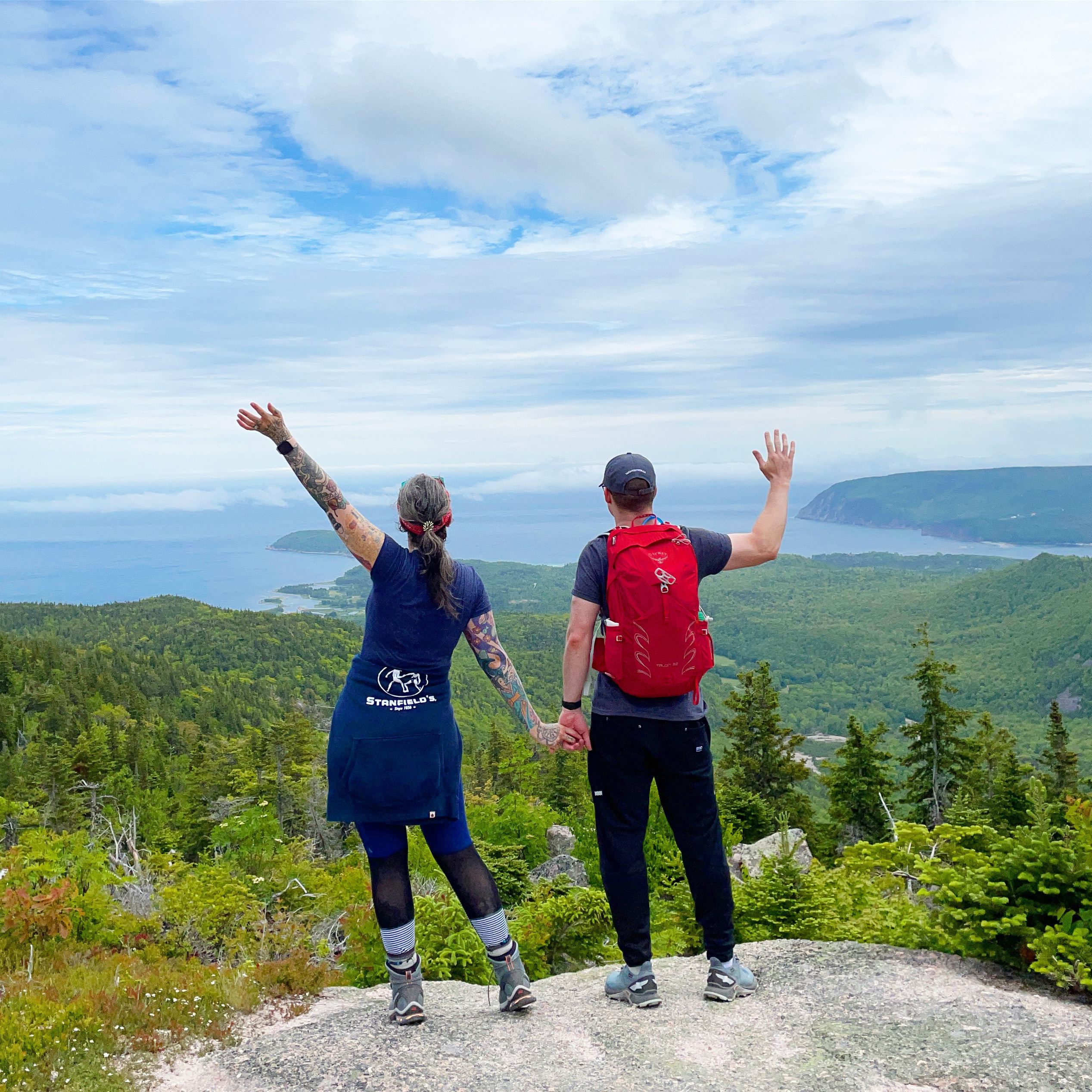 A man and woman hold hands and wave out to the ocean as they stand on top of a hiking summit surrounded by trees below