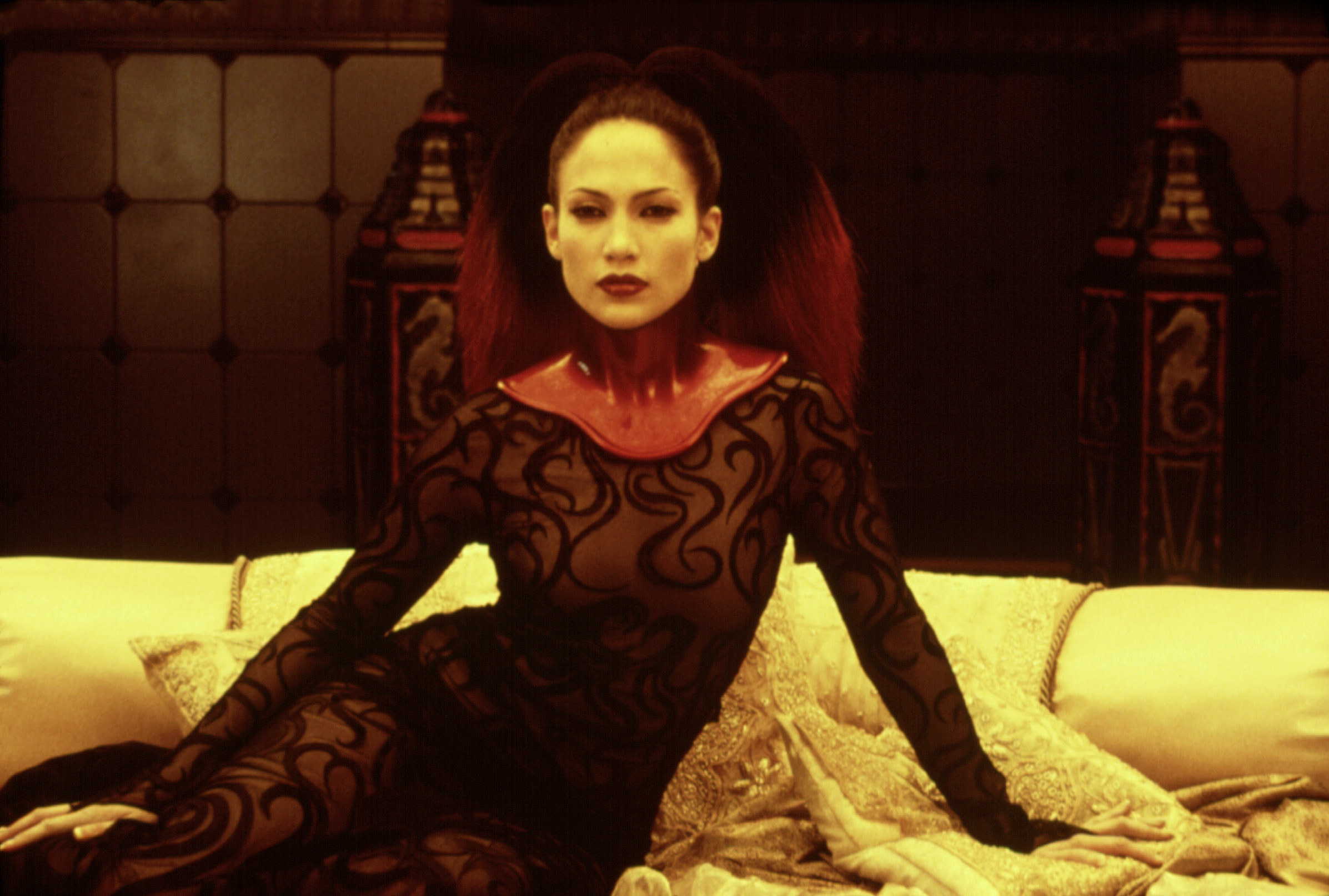 Jennifer Lopez in an ornate bodysuit and red leather collar lounging on a bed
