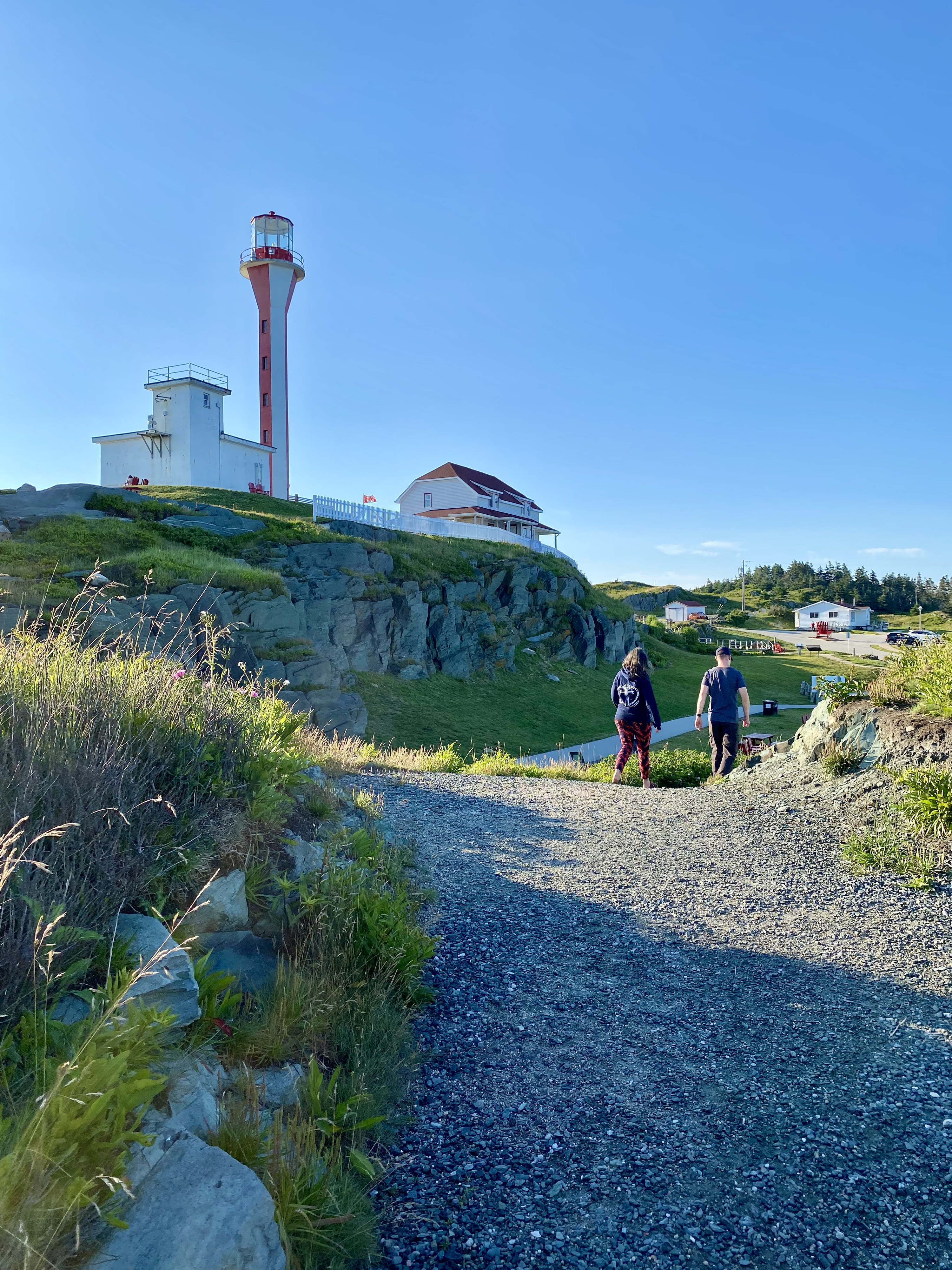 A man and woman walk along a gravel path with a lighthouse in the background