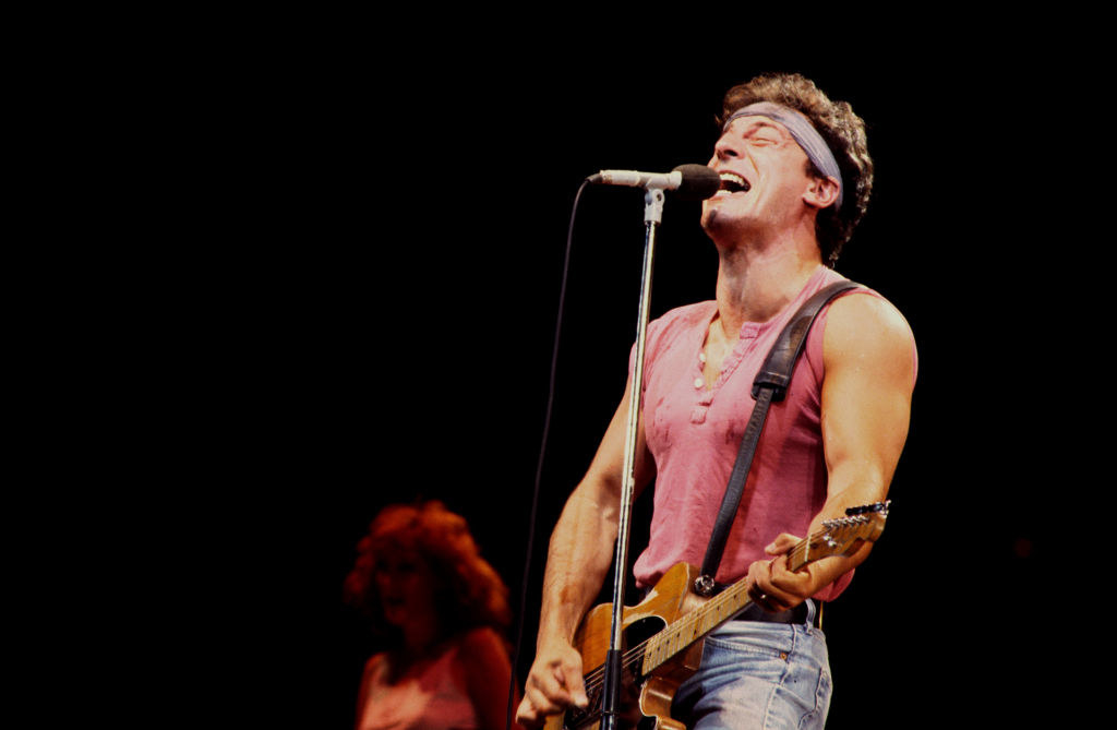 Bruce Springsteen singing onstage in a shirt and jeans and bandana and playing guitar
