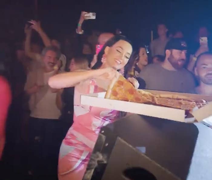 Katy reaches into a box of pizza while standing behind a DJ