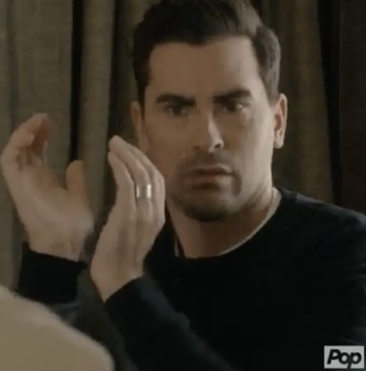 A person with a serious expression claps their hands, wearing a dark sweater, in a scene from a TV show