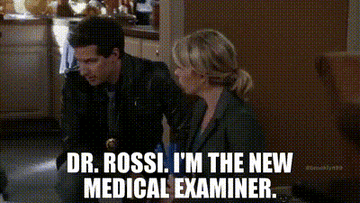 A blonde woman with a grey sportcoat kneels next to Jake Peralta as white text appears reading &quot;Dr. Rossi, I&#x27;m the new medical examiner&quot;