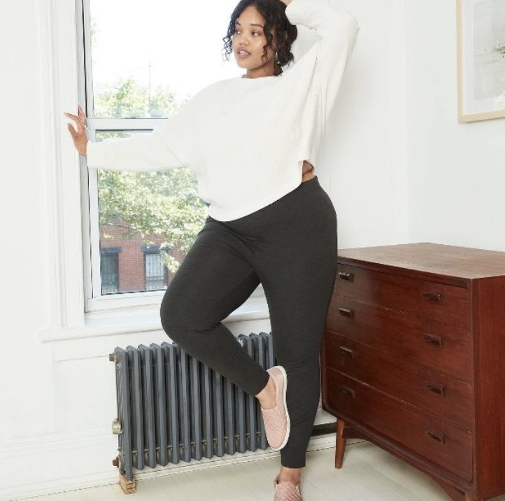 The model wears the dark leggings while posing in an apartment