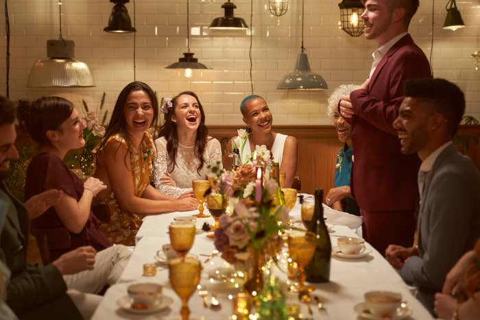 Friends laugh at a table during a wedding reception. A man stands at the center of the table.