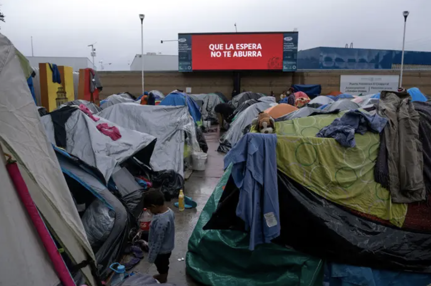 An immigrant child stands amid tents at an improvised camp near a screen reading &quot;May the wait not bore you&quot; in Tijuana, Mexico. 