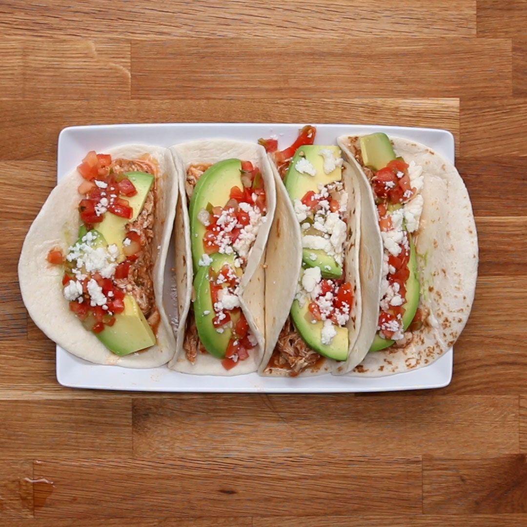 Four soft tacos with shredded chicken, avocado slices, diced tomatoes, and crumbled cheese on a white plate, displayed on a wooden table