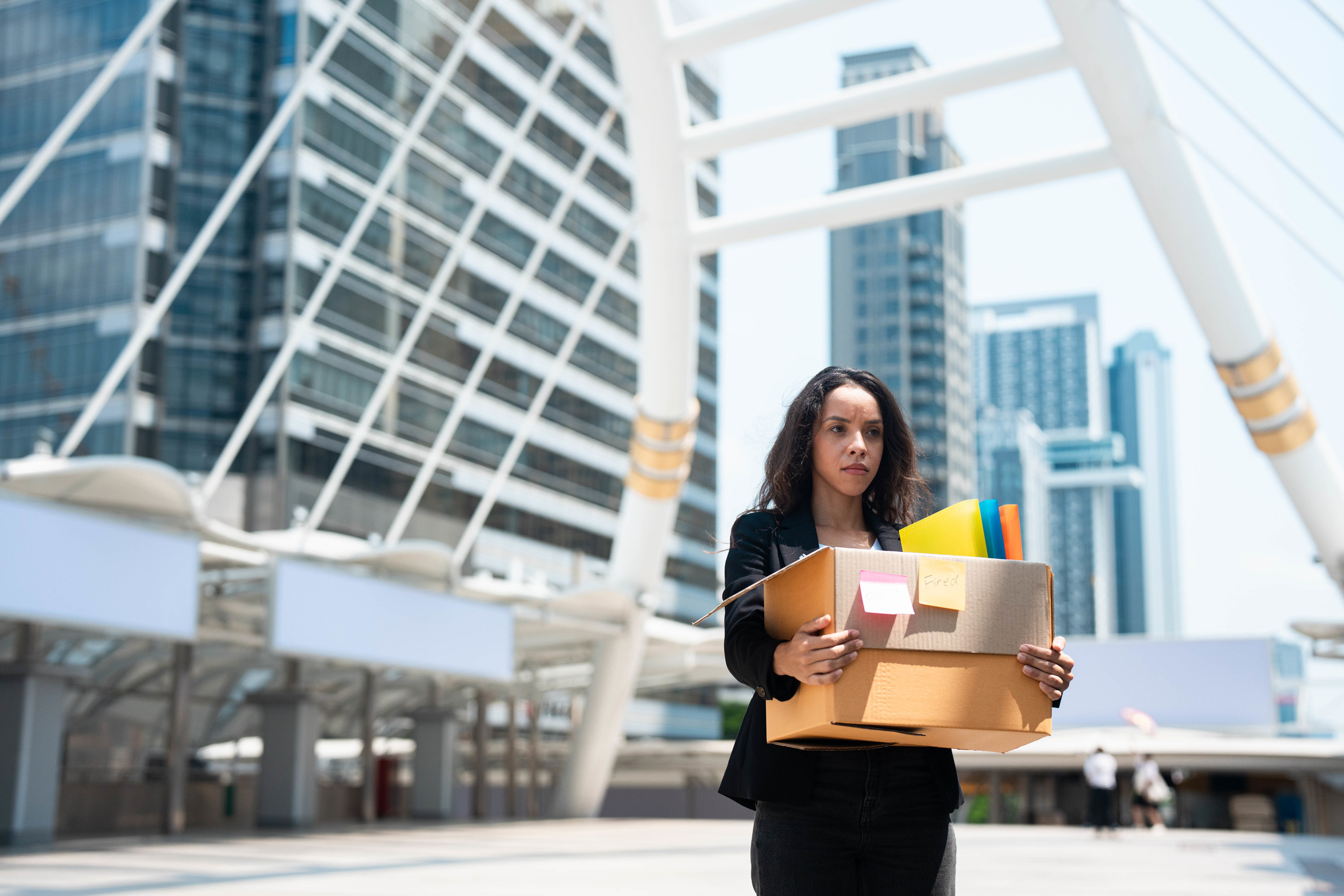 woman walking away from an office building with a cardboard box after being laid off