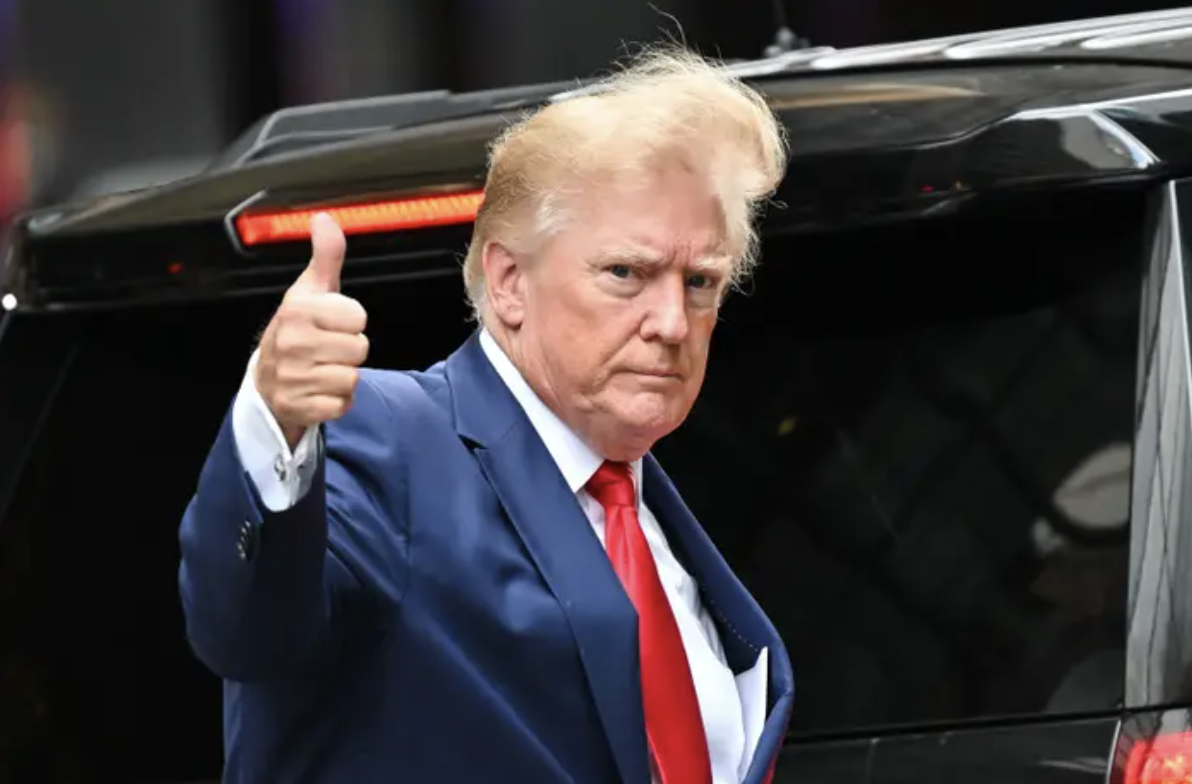 Trump wearing a suit with a red tie giving a thumbs up