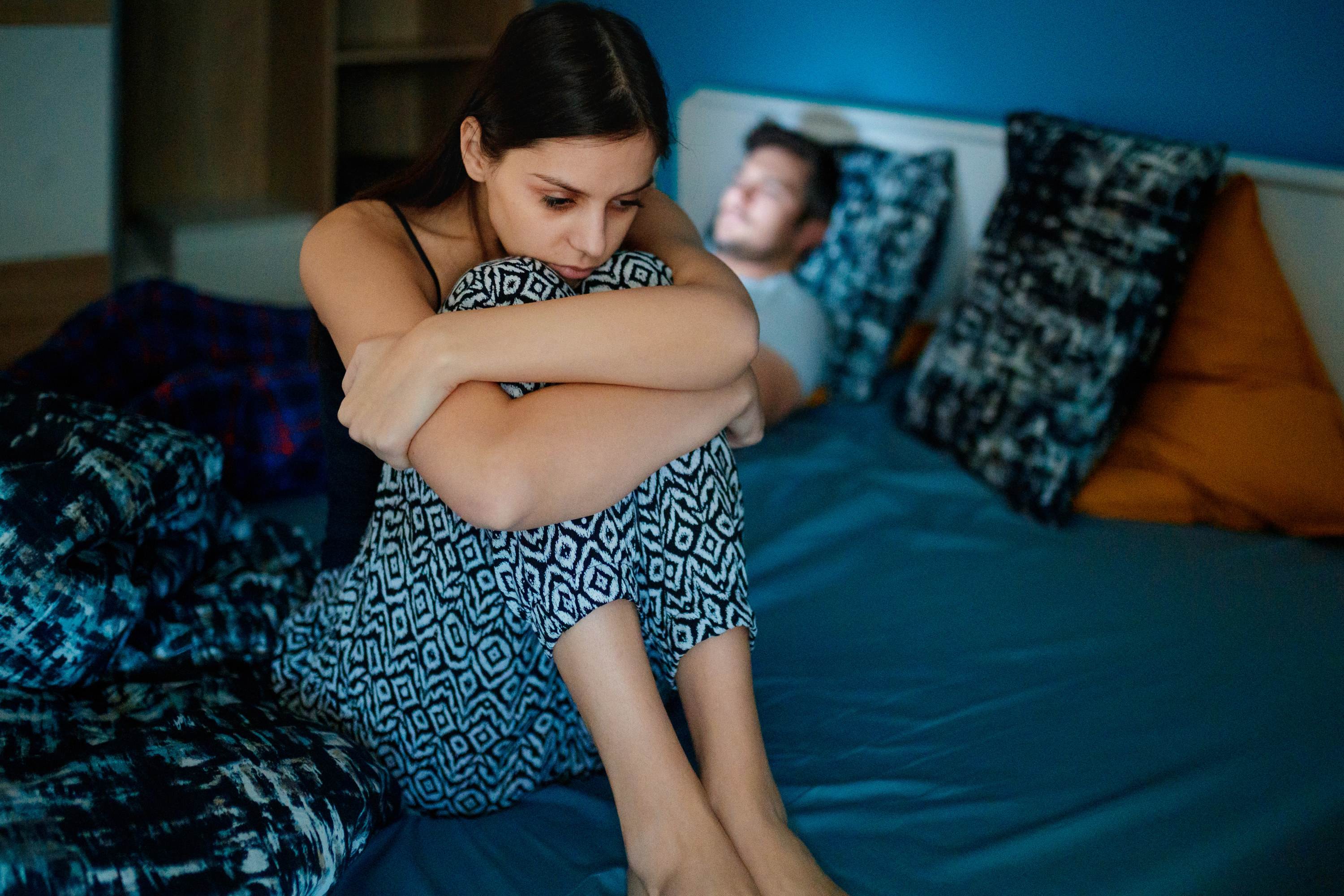 Woman sitting up in bed and hugging her knees with a man lying in bed behind her