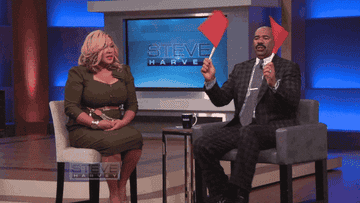 Steve Harvey dancing on his show as he holds two red flags and sits next to a lady talking