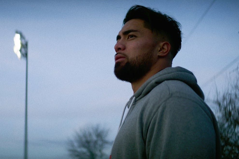 Manti Te&#x27;O wears a grey hoodie and stares off into the distance, he&#x27;s outside, it&#x27;s dusk, and the sky is gray