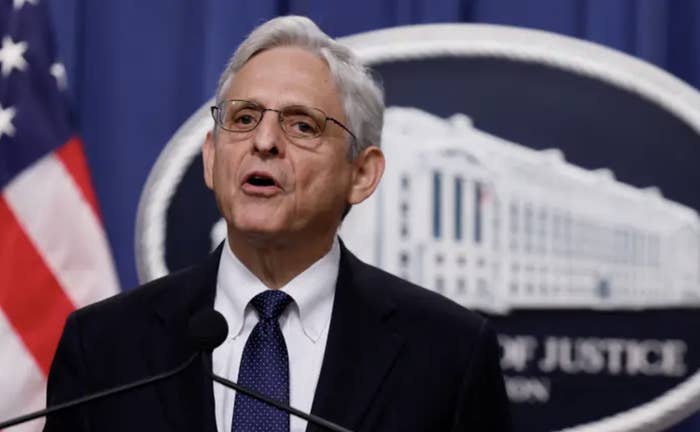 Merrick Garland wears a suit and glasses and stands at a podium with a department of justice sign and american flag in the background