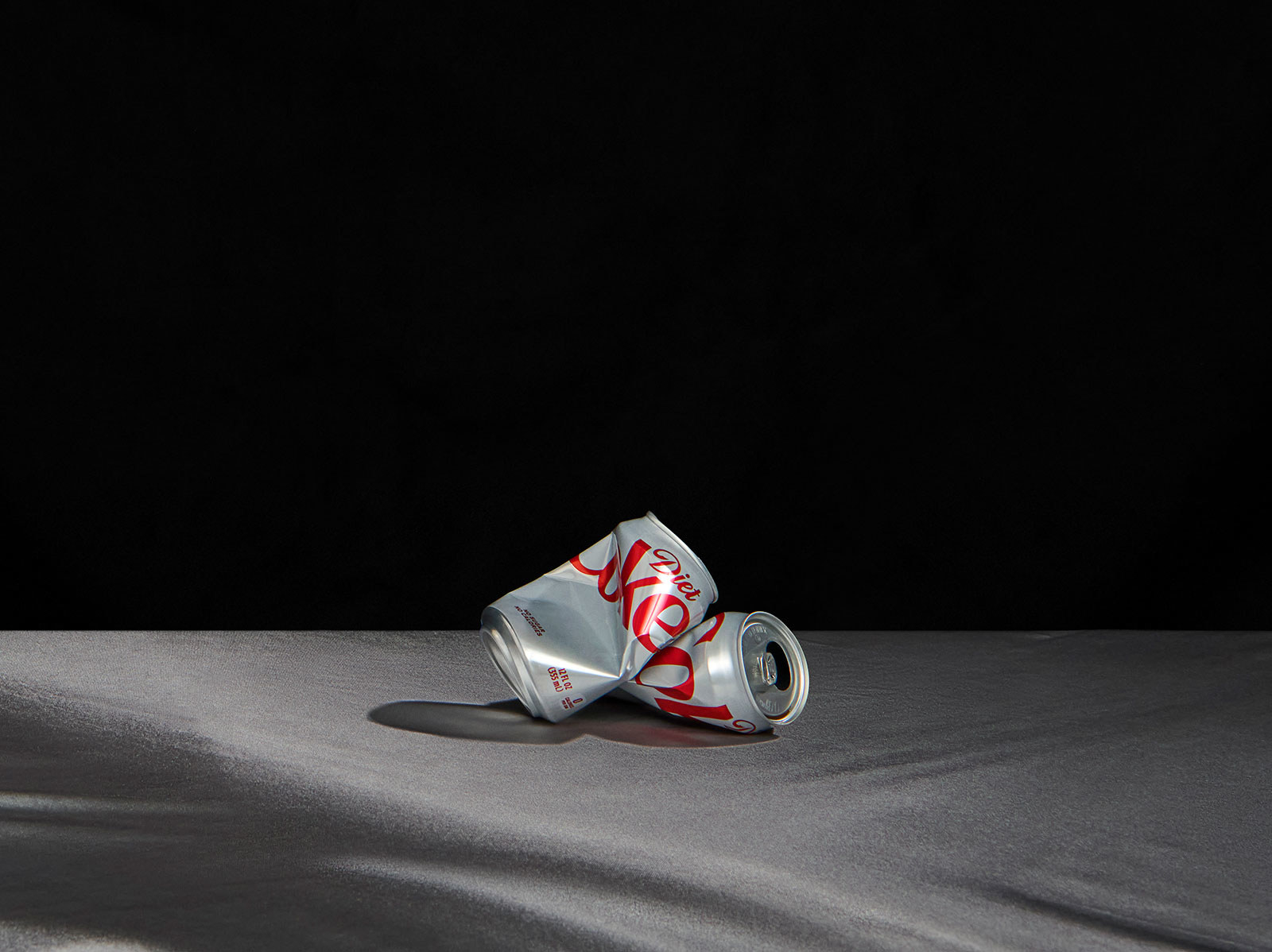 Two crumpled Diet Coke cans on top of each other on a black blanket in a dark photo studio