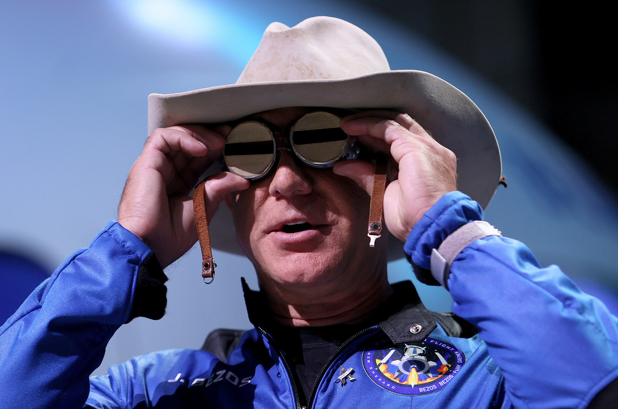 Jeff Bezos wearing a blue space suit and cowboy hat holds the aviation glasses that belonged to Amelia Earhart to his eyes