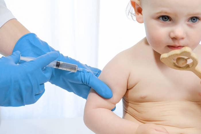 A baby sits as a pair of gloved hands injects a syringe into their arm
