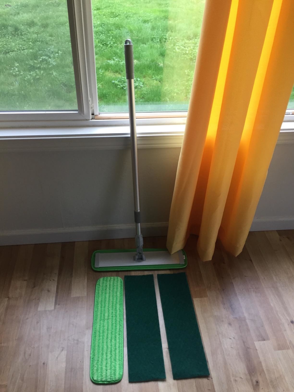 Reviewer&#x27;s photo of the mop resting against window on hardwood floor with extra microfiber and two scrubbing pad attachment heads laid out in front