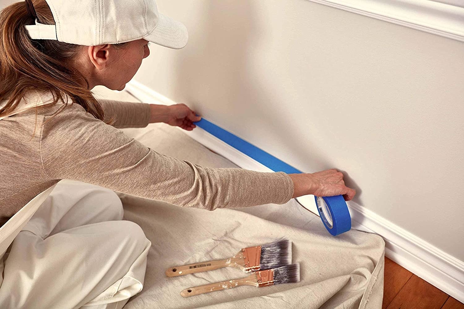 a model applies painter&#x27;s tape to a wall while standing atop the canvas drop cloth