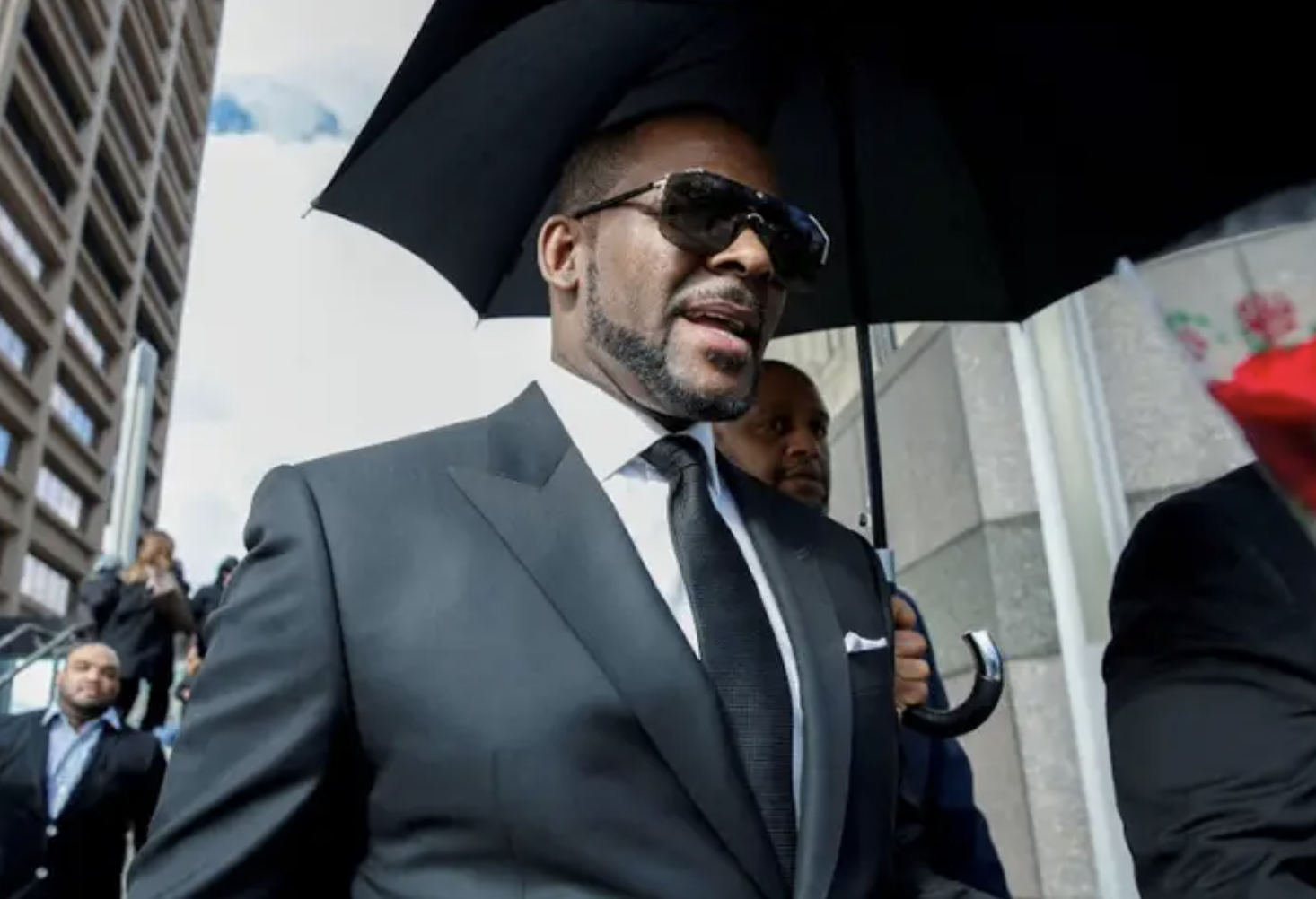 r. kelly walks outside in a suit and sunglasses. someone holds an umbrella for him