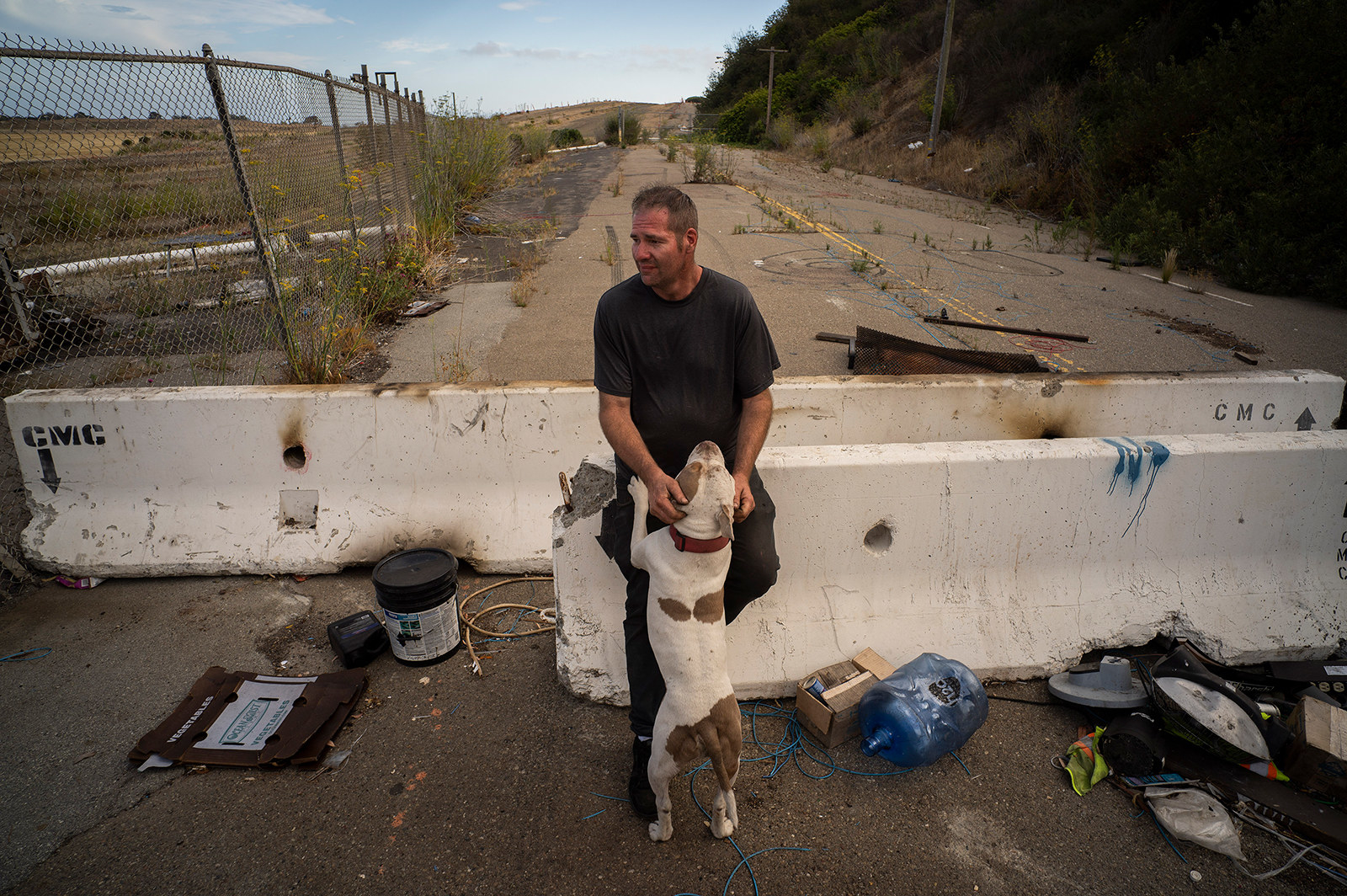 A man sits on a concrete barrier surrounded by debris, while his dog jumps up on him