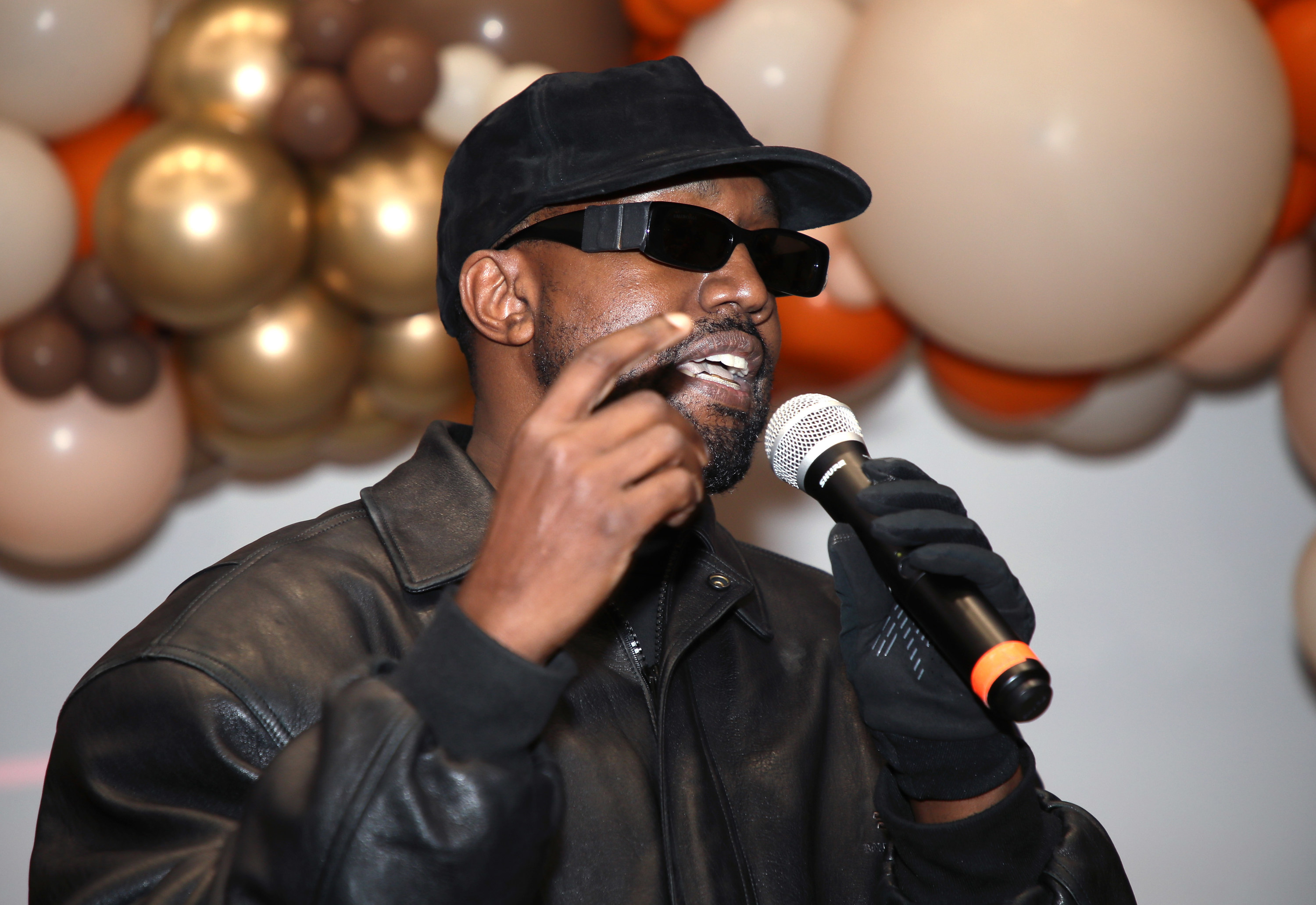 Ye wearing a cap and sunglasses and speaking into a microphone