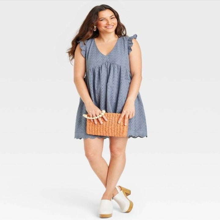 Model wearing slate blue dress, white clogs and holding a rattan clutch