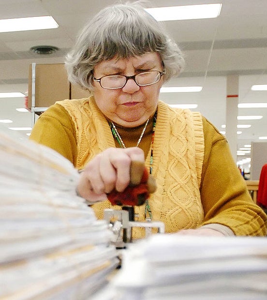 A woman with a yellow vest over a yellow sweater, with short grey hair and reading glasses stamping tax forms