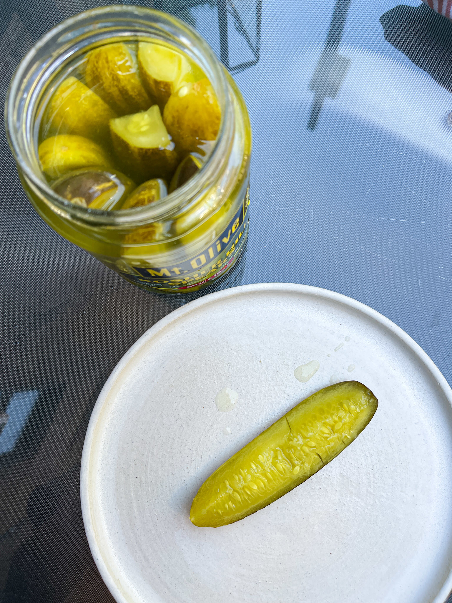 Open jar of pickles with one pickle on a paper plate, on a glass table