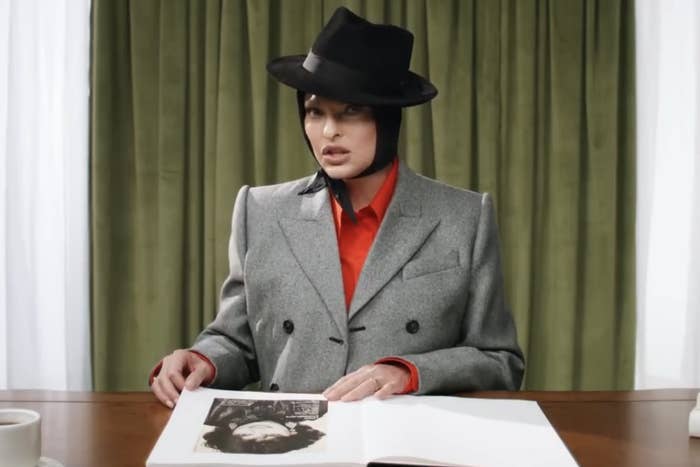 Linda sitting at a table with a coffee table book open in front of her and wearing a hat with attached scarf tied under her chin