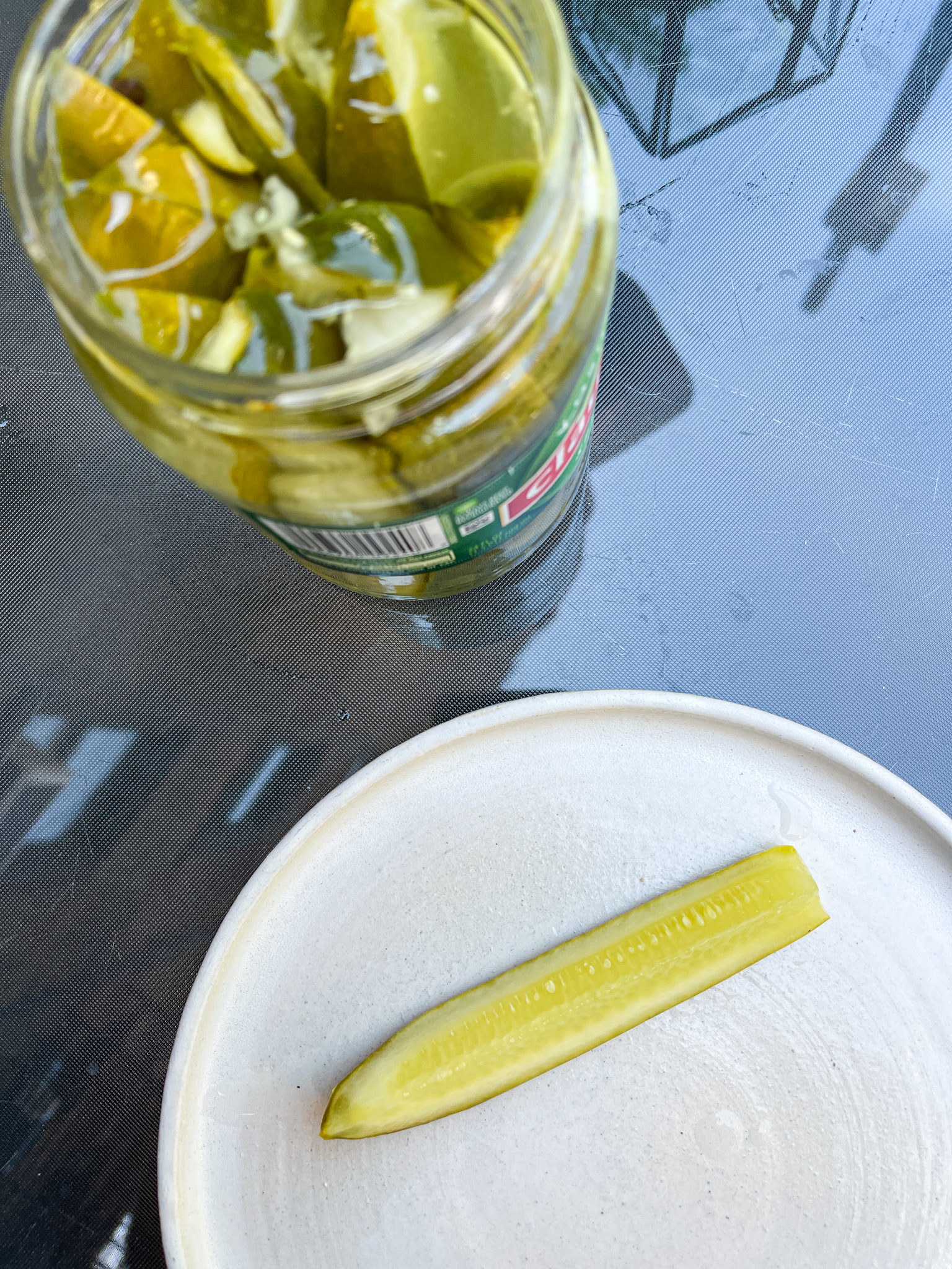 Top view of an open pickle jar and a single pickle slice on a plate on a glass tabletop