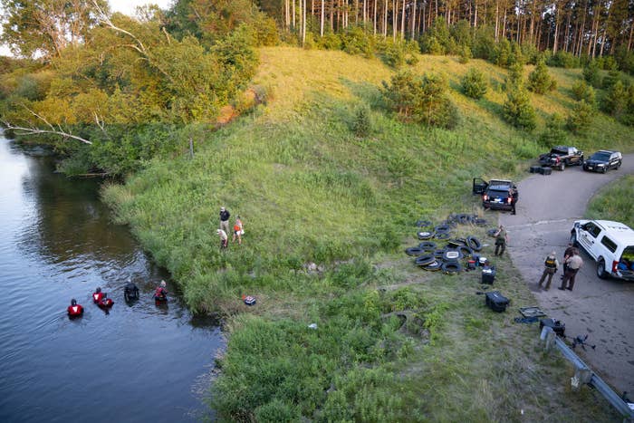 An overhead view of a grassy area between a river, where people wearing bright vests dig in the water, and a small road with parked law enforcement vehicles and officers