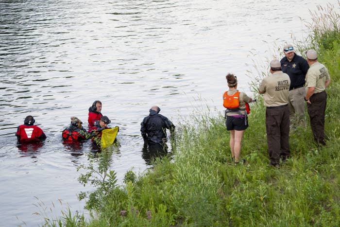 Five people stand in a river near four others just offshore in a grassy area