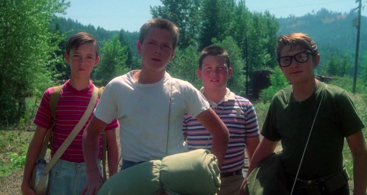 The four boys from Stand by Me stand in a row in front of a cluster of trees