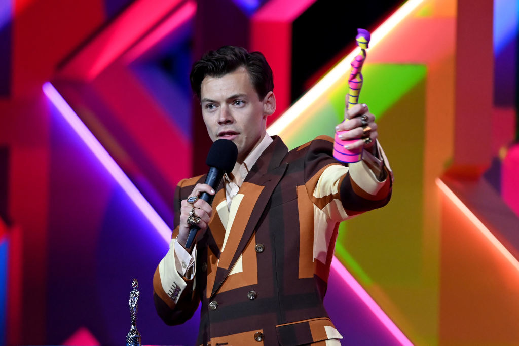 Harry in a colorful jacket and speaking into a microphone onstage while holding an award