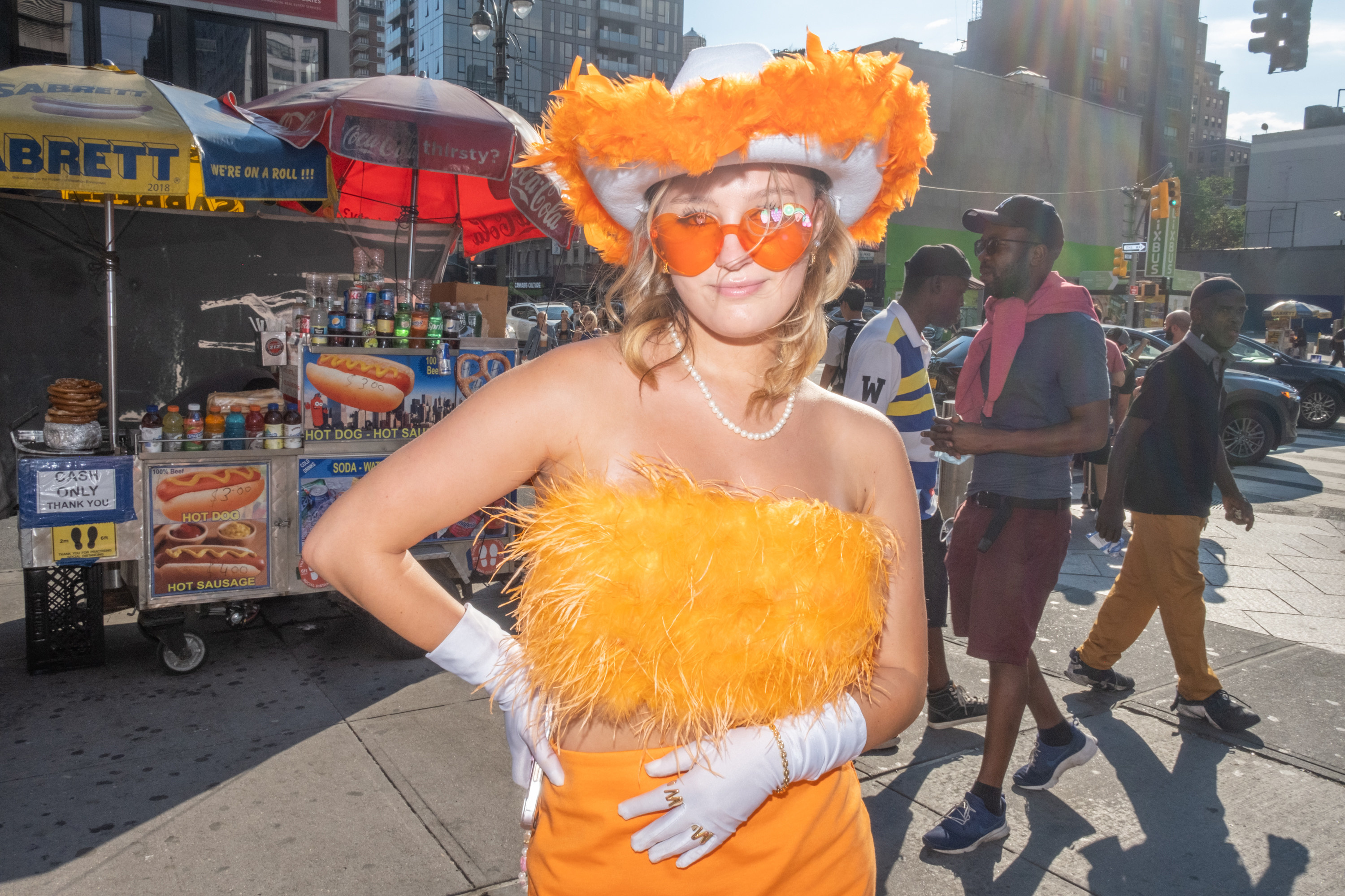 A woman with a vibrant orange outfit, heart-shaped sunglasses, and rings that spell &quot;MW&quot; stands in front of a hot dog cart