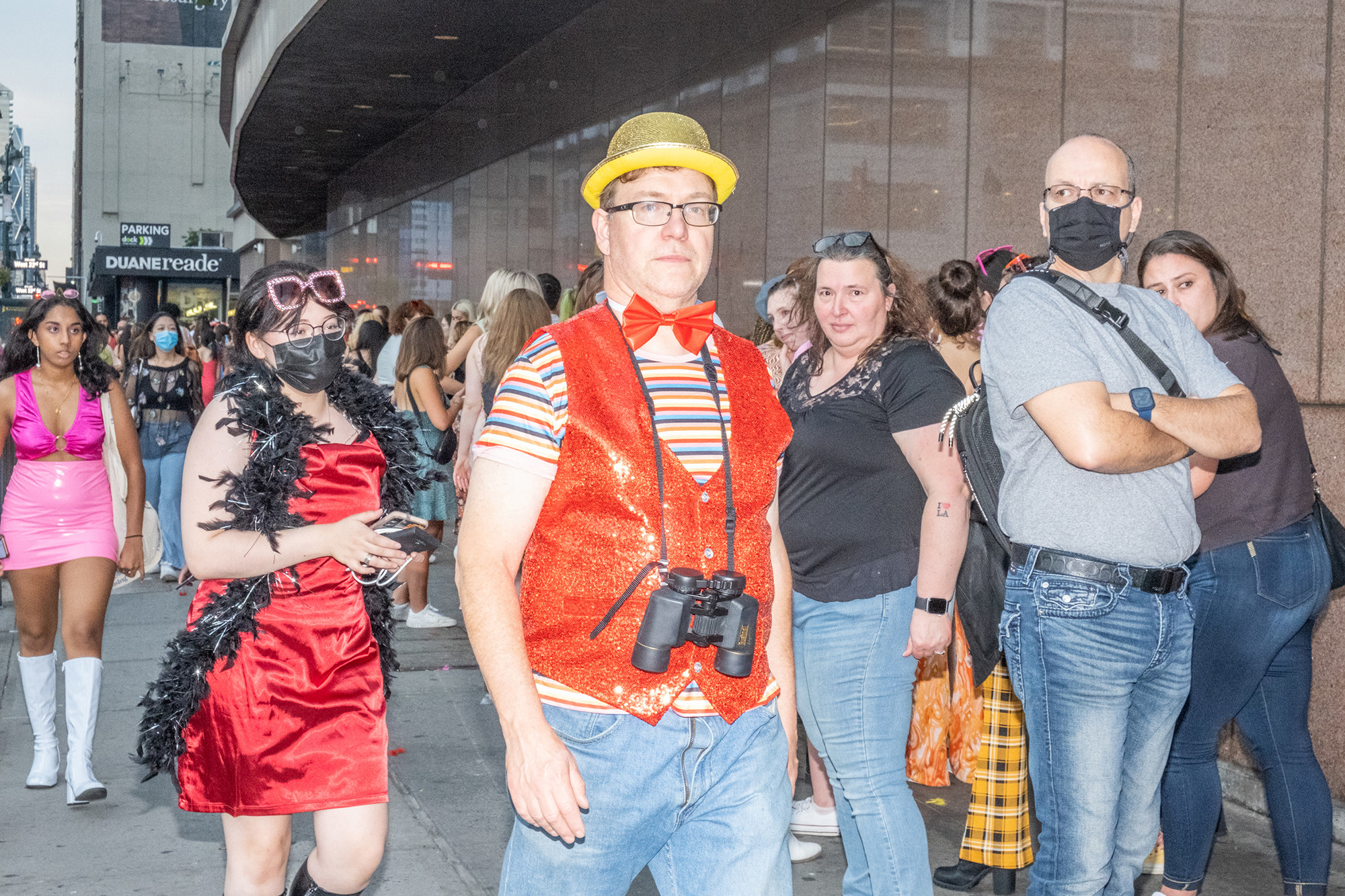 A man with a tiny yellow bowler hat, sparkly orange vest, and binoculars waits in line outside Madison Square Garden