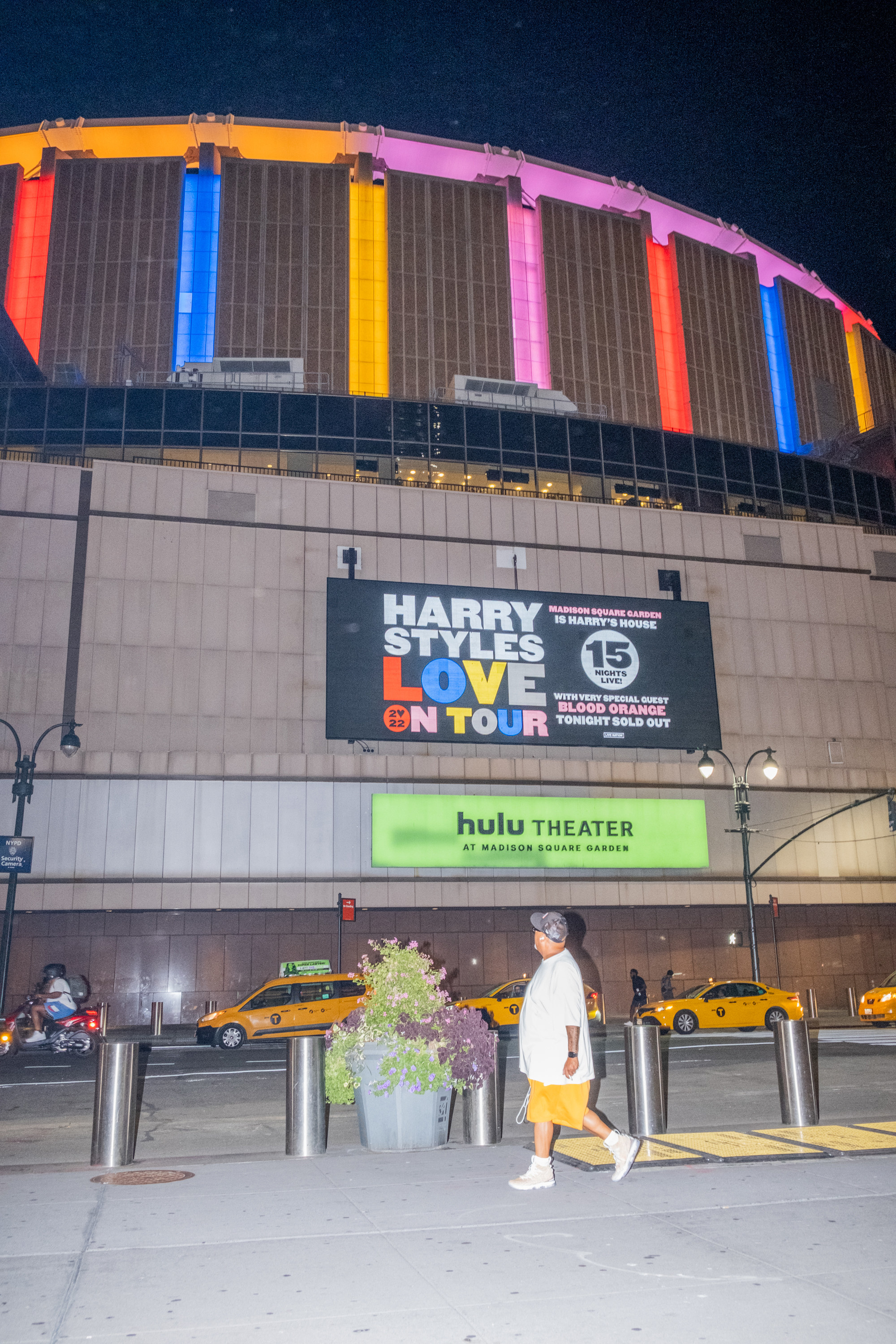 A man walks in front of Madison Square Garden at night where a sign advertising Harry Styles&#x27; tour is visible