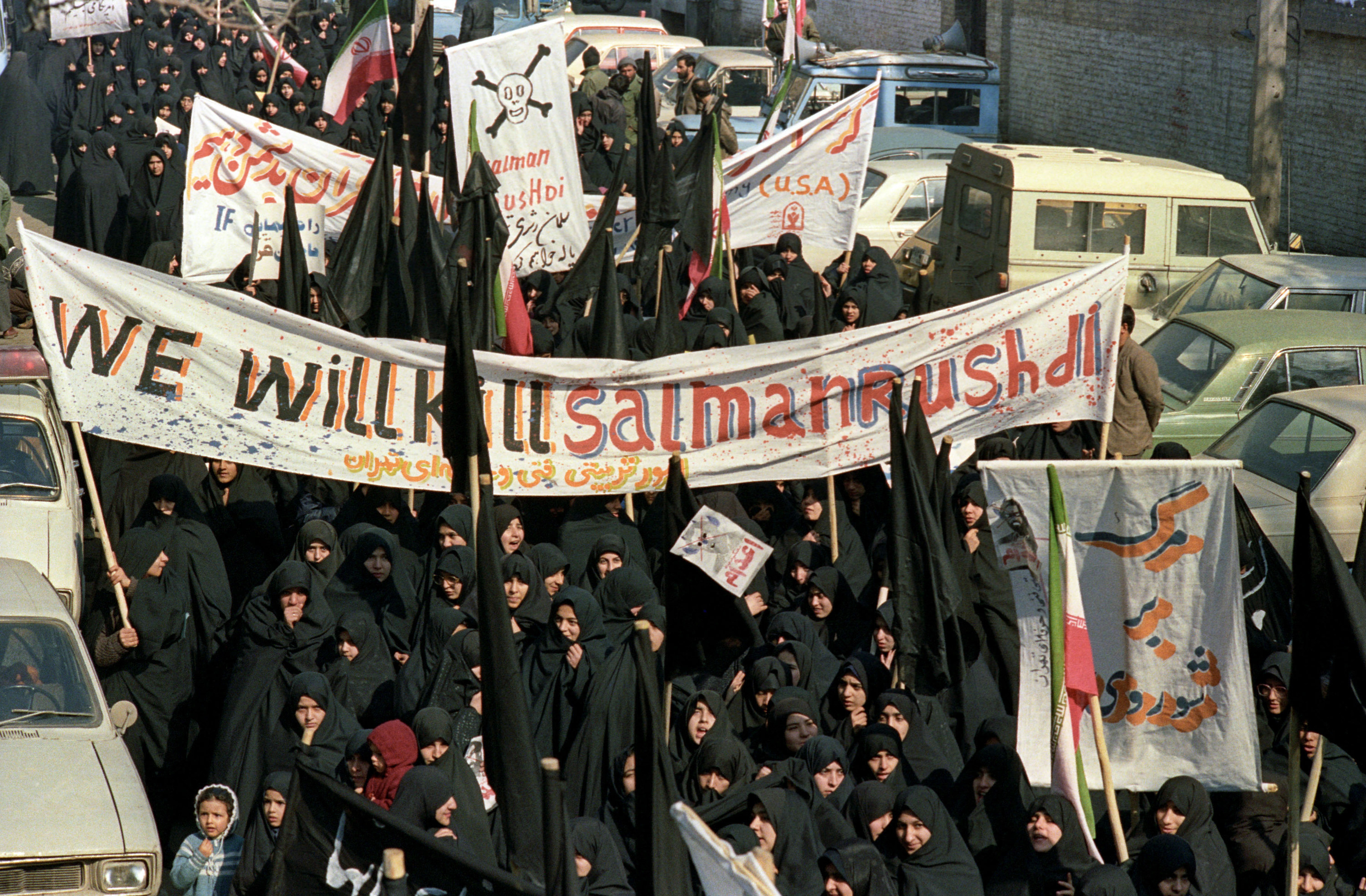 A crowd of people dressed in black protest and hold signs reading &quot;we will kill salman rushdie&quot;