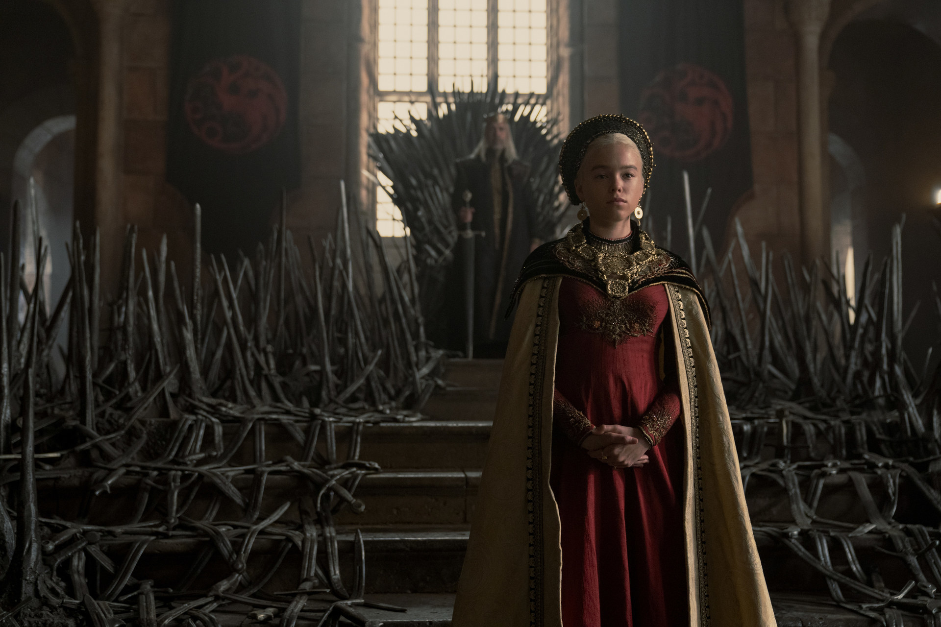 Rhaanyra standing in front of the throne on which Viserys sits