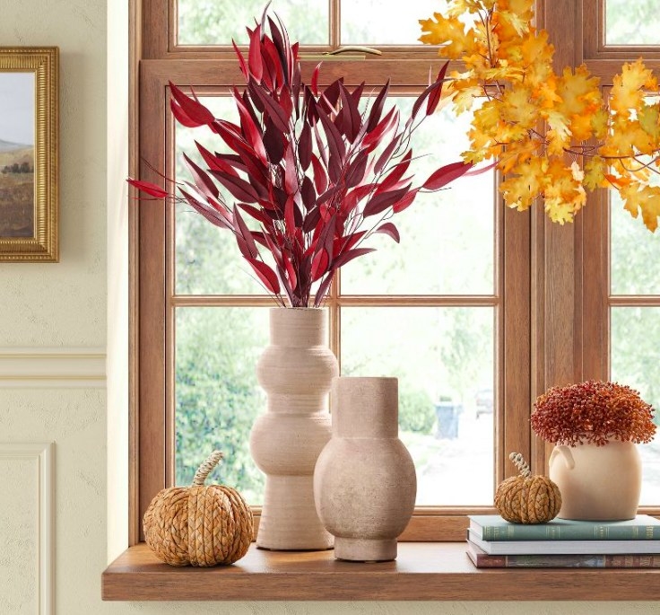 Ceramic vases, one filled with red stems, on a window sill styled with fall decorative items