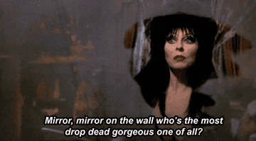 Cassandra Peterson with red lipstick and big black hair saying &quot;Mirror, mirror on the wall who&#x27;s the most drop dead gorgeous one of all&quot;