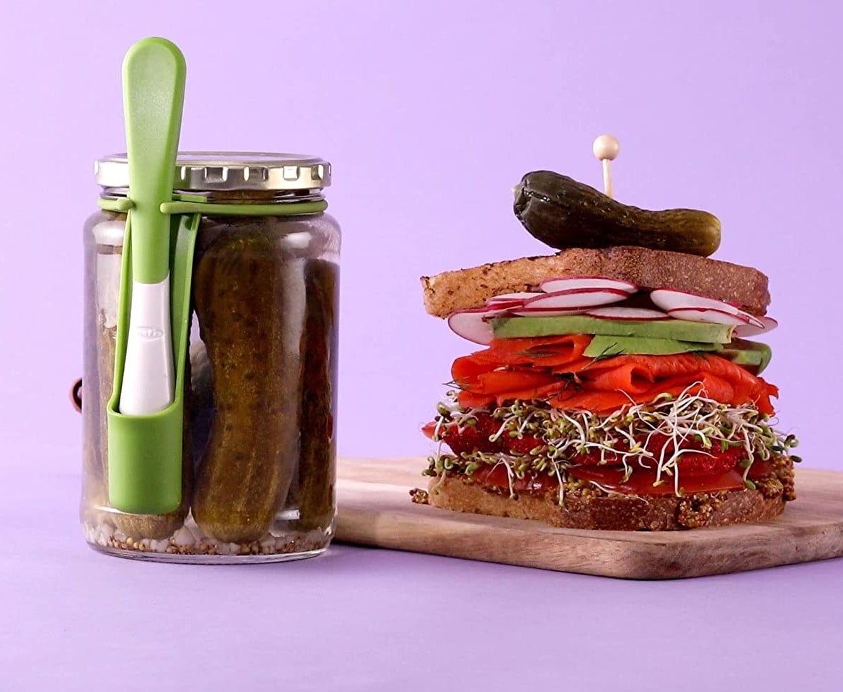 The condiment fork attached to a pickle jar beside a sandwich