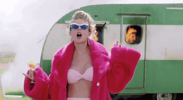 Taylor Swift walking away from a burning caravan holding a cocktail and wearing a pink fluffy coat