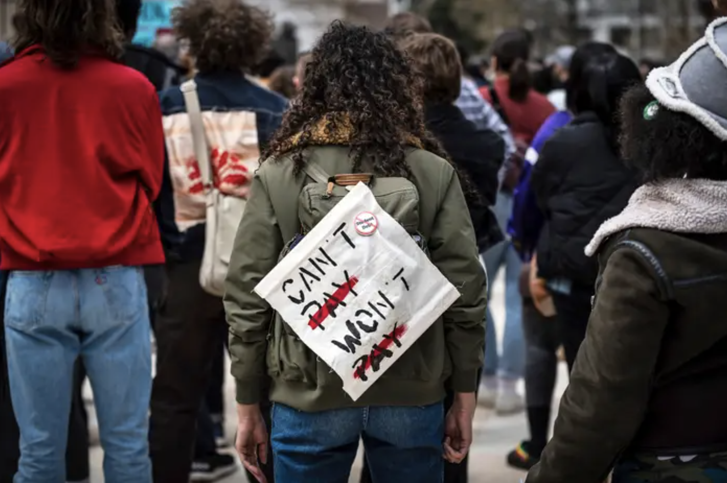 A person in a green jacket and curly hair stands in a crowd with their back to the camera. on their back is a canvas sign that says &quot;can&#x27;t pay, won&#x27;t pay.&quot;