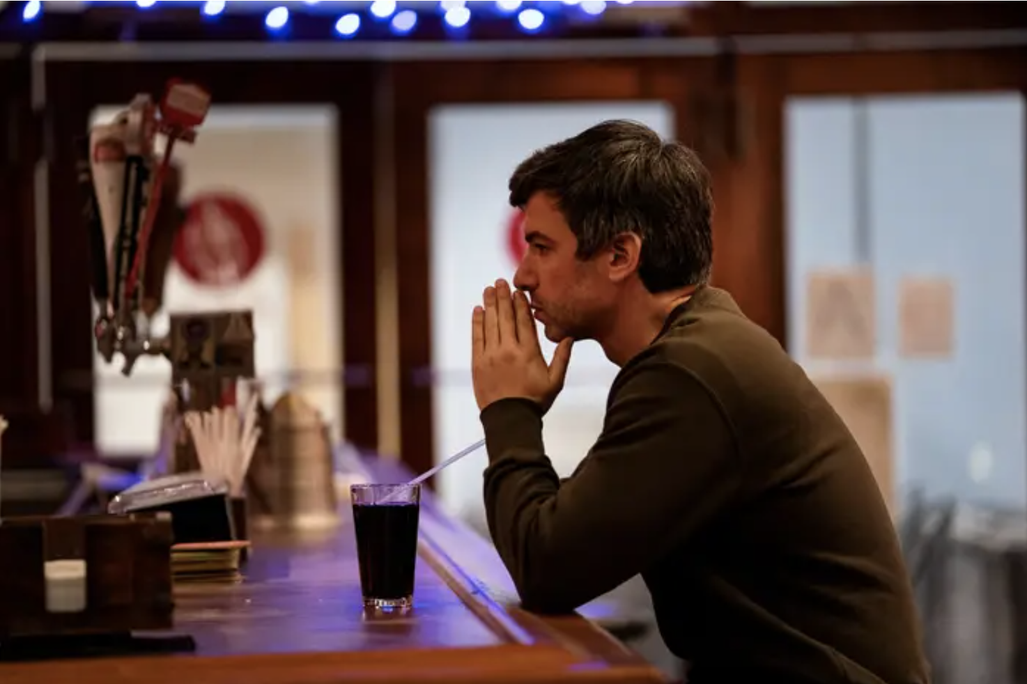 nathan fielder wears a brown sweater and sits at a wood-furnished bar looking pensive 