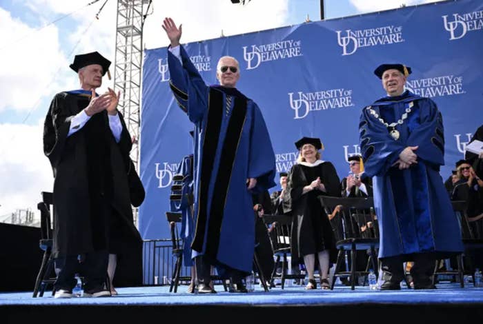 biden stands onstage in black sunglasses and a blue graduation robe waving to a crowd. other people in robes are on his left and right