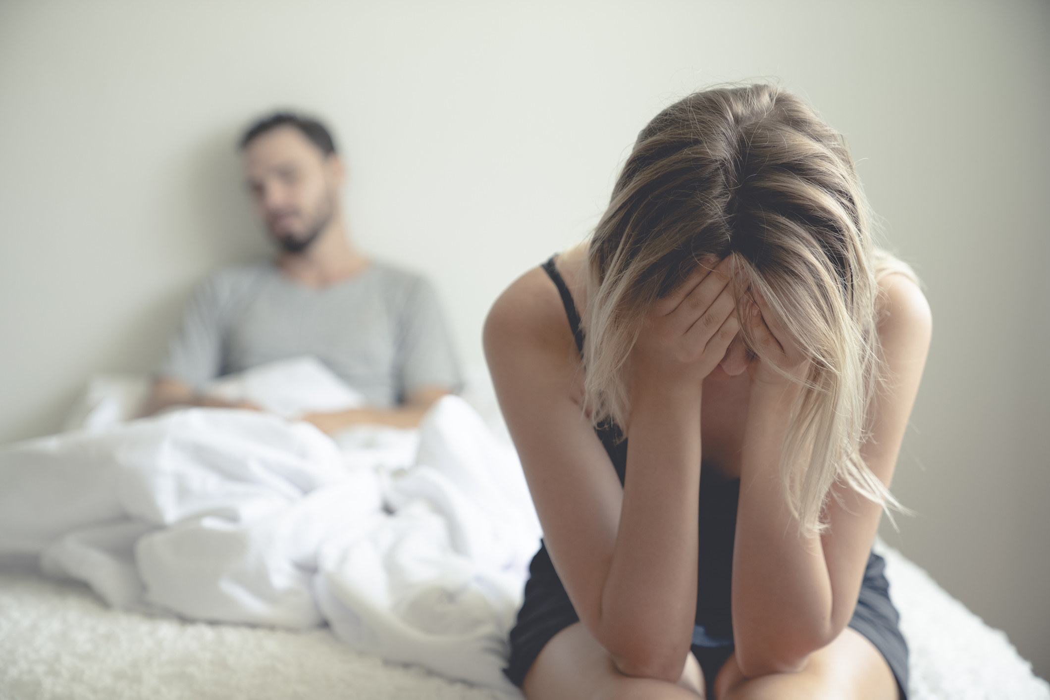 Woman sitting on a bed with her head in her hands as a man sits up in bed behind her