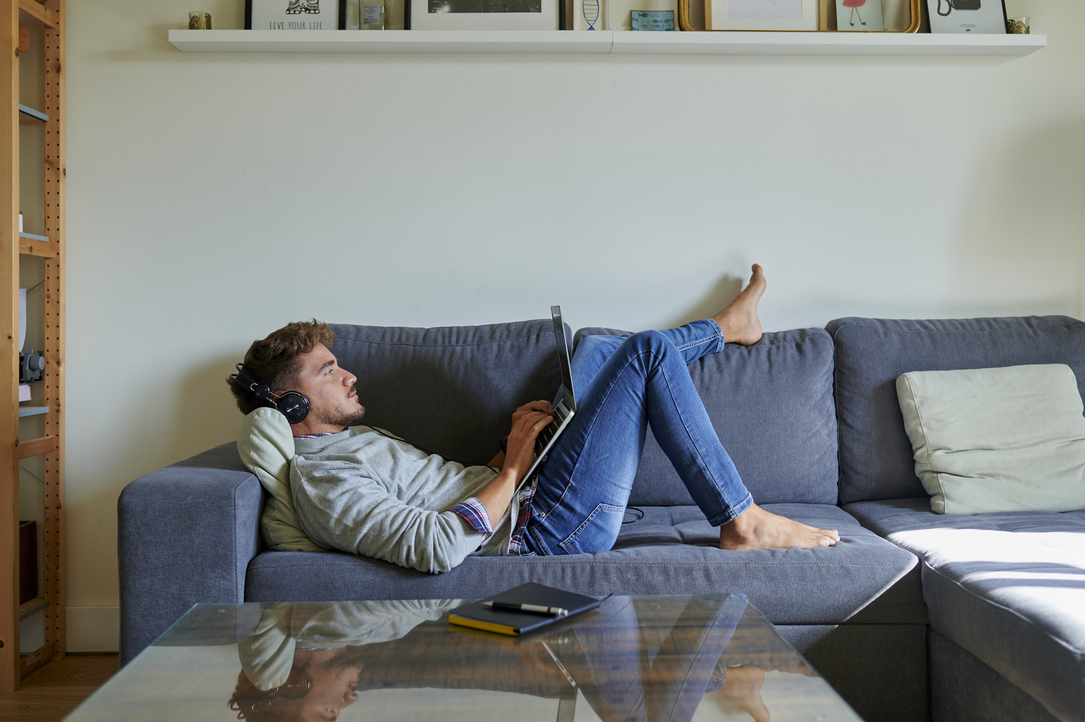 Man lying on a couch working on a laptop and wearing headphones