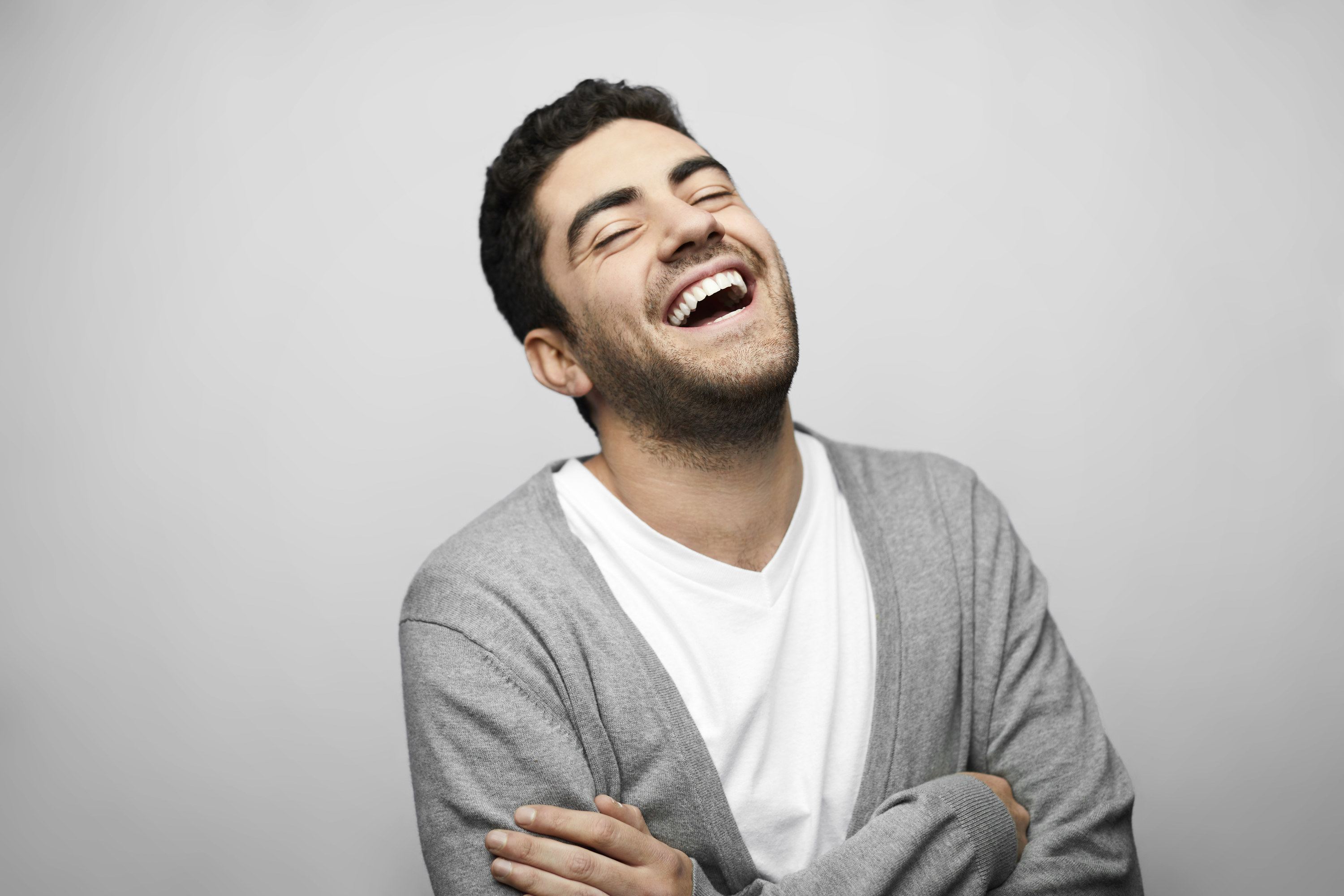 Latin American young man laughing against gray background. Cheerful male is with arms crossed. He is wearing casual.