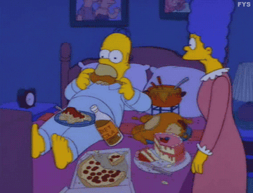 荷马·辛普森躺在床上,吃一个鸡腿,包围着盘子的食物,一个披萨,蛋糕和芝士火锅锅